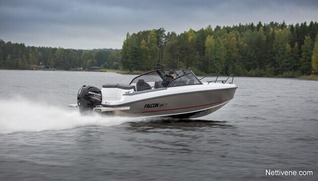 A white Falcon 590 boat speeds across a calm lake, leaving a wake, surrounded by green trees under a cloudy sky.